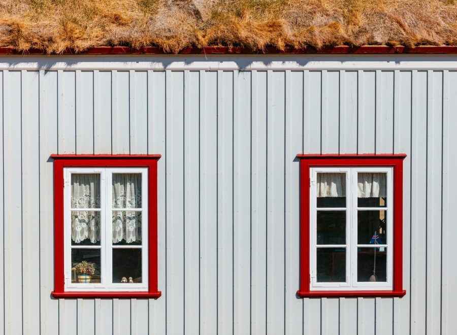 Vit panelfasad med två spröjsade fönster i röda fönsterfoder under torvtak